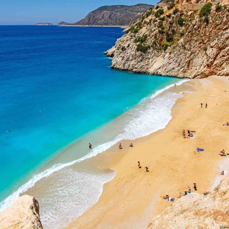 Strand mit Felsen und Menschen am blauem Wasser.