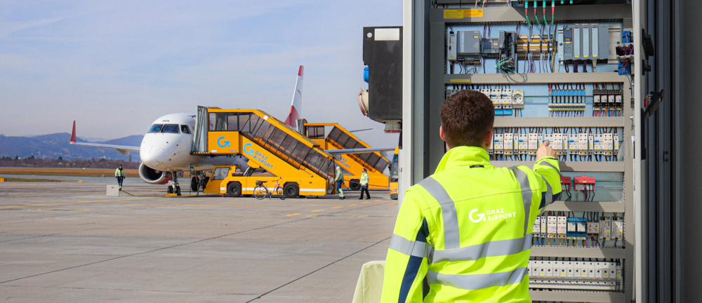 Schichtelektriker:in vor dem offen Schaltschrank am Vorfeld mit Flugzeug im Hintergrund